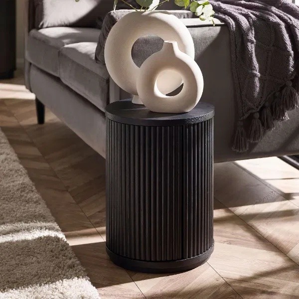 Black fluted side table with a round top and door, featuring a textured finish, placed beside a gray sofa and adorned with white decorative sculptures.
