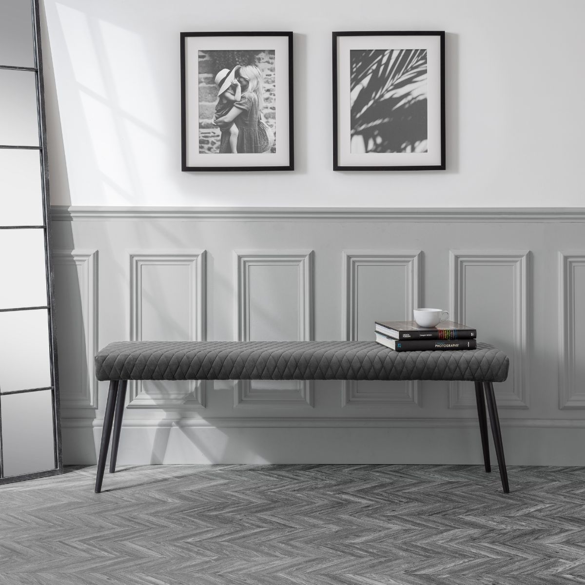 Grey upholstered low bench with a quilted design, featuring black tapered legs, positioned against a light wall with two framed pictures above.