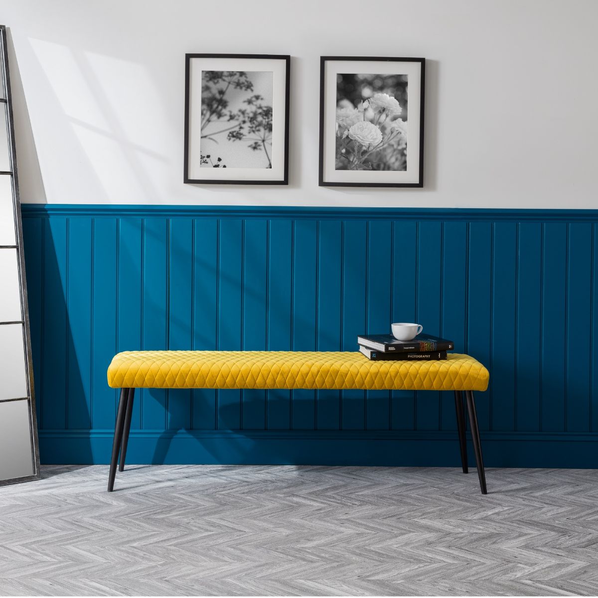 Mustard yellow upholstered low bench with a quilted design and sleek black legs, placed against a blue paneled wall, with framed artwork above and a book on top.