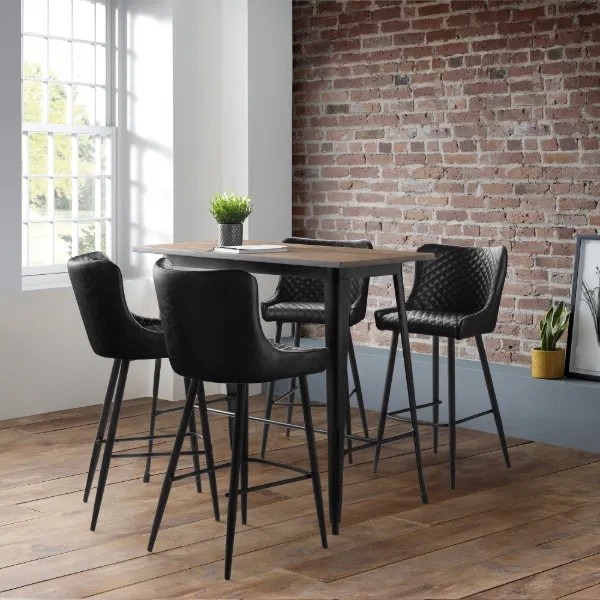Black faux leather barstool with a quilted backrest and sleek metal legs, positioned around a wooden table in a modern, stylish interior with exposed brick walls.