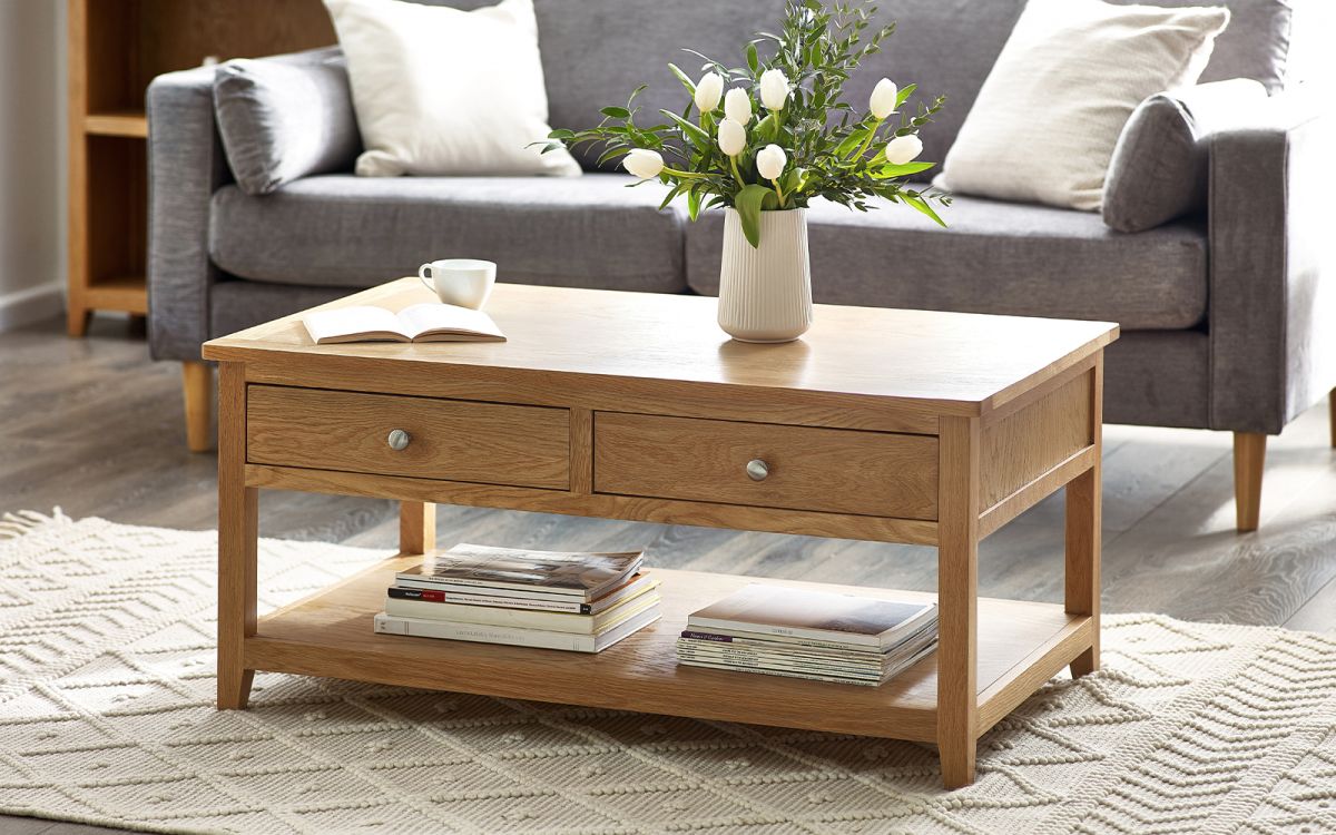 Brown light wood coffee table featuring two drawers and an open shelf, adorned with a vase of flowers, a cup, and a book, set in a cozy living room.