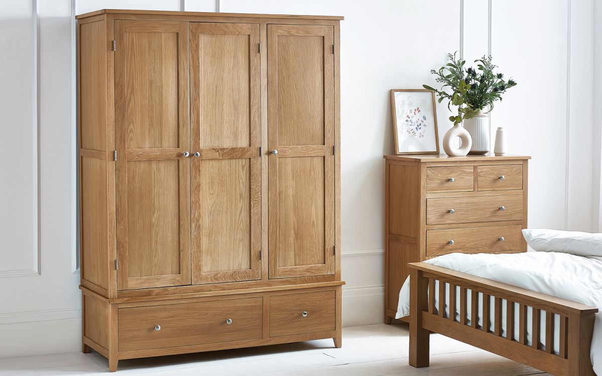 Light wood wardrobe with three doors, featuring a smooth finish, silver knobs, and two lower drawers, set against a neutral wall and accompanied by a bedside table and decorative items.