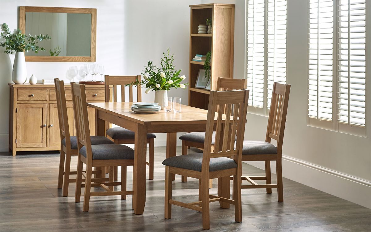 Light wood extending dining table with a smooth finish, surrounded by six wooden chairs featuring grey upholstered seats, set in a bright, modern dining room with natural light.
