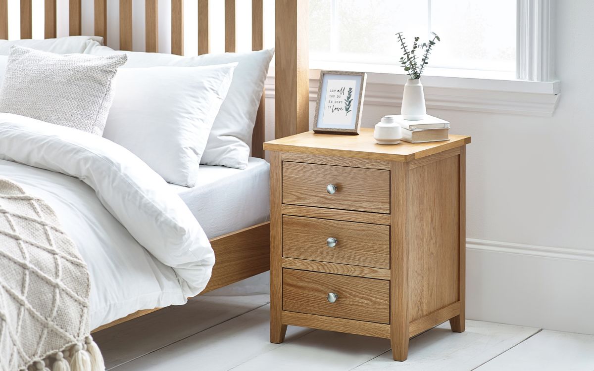 Light wood bedside table with three drawers, silver knobs, and a smooth top, positioned beside a bed with white bedding and a small decorative vase on top.