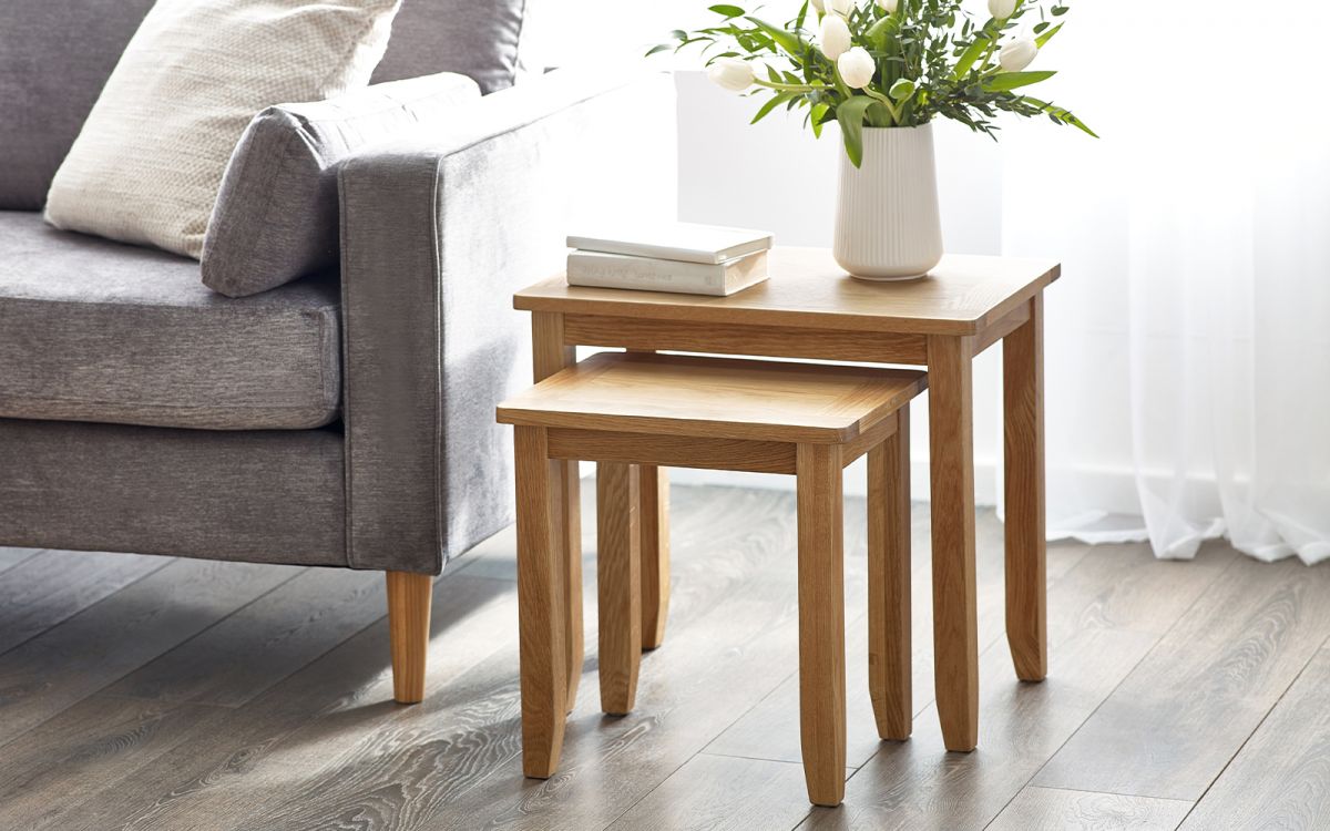 Light wood nest of two tables with clean lines, featuring a smooth surface and tapered legs, placed beside a grey sofa and adorned with a white vase of flowers and books.