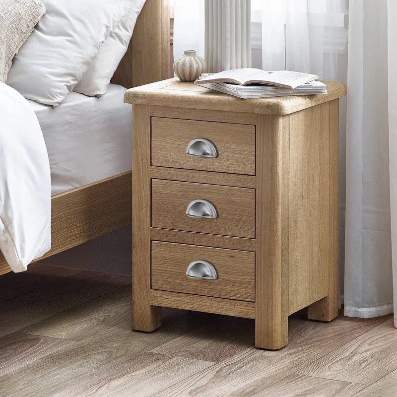 Three-drawer bedside table in limed oak finish, featuring rounded edges, silver metal handles, and a smooth top, positioned next to a bed with white bedding and a book on top.