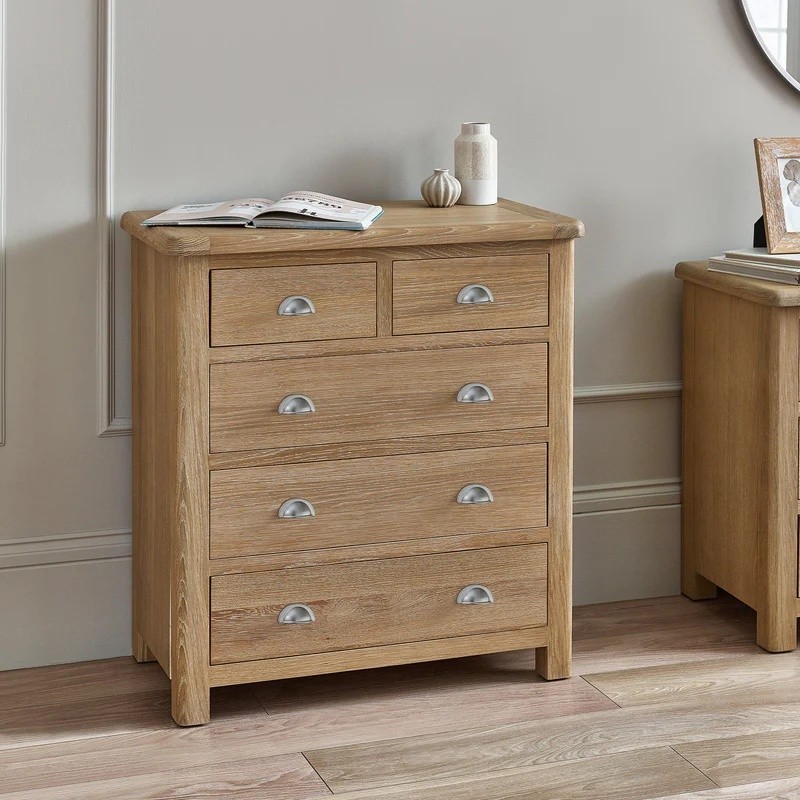 Limed oak chest with three small and two large drawers, featuring curved edges and silver handles, set against a light wall and wooden floor.