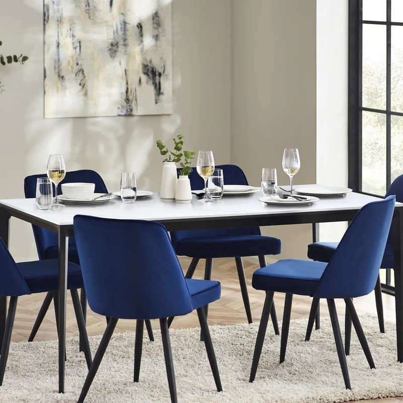 Rectangular dining table with a white marble top and black metal legs, surrounded by six blue upholstered chairs, set with glasses and plates in a bright, modern room.