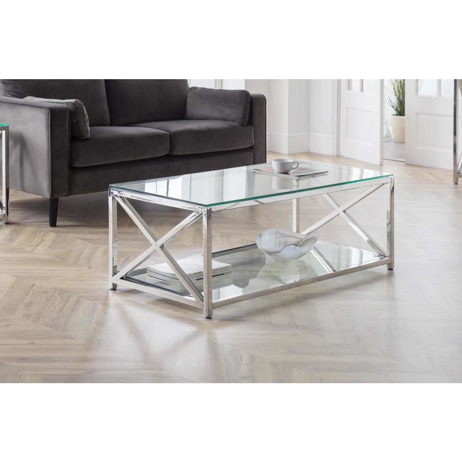 Miami Coffee Table features a sleek glass top and silver metal frame with an X-shaped design, set on a wooden floor, accompanied by a gray sofa and decorative items.