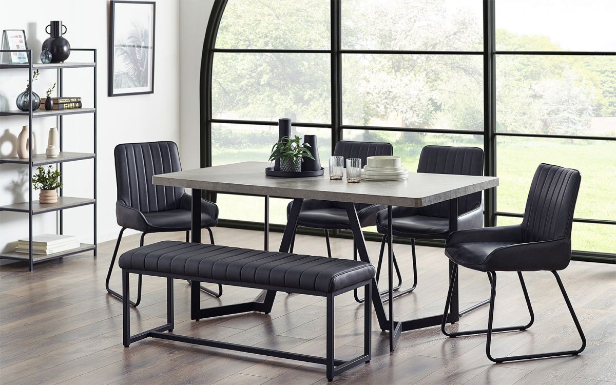 Grey concrete effect dining table with a sleek design, accompanied by black upholstered chairs and a matching bench, set against a large window with a view of greenery.
