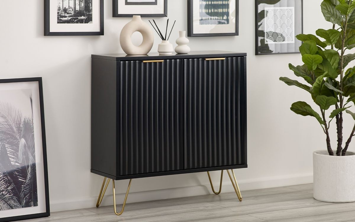 Matt black compact sideboard with ribbed doors, gold handles, and tapered metal legs, accompanied by decorative vases and framed artwork against a white wall.