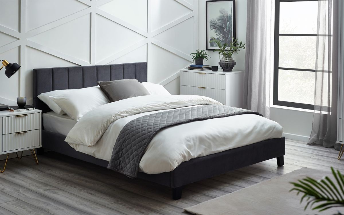 Grey velvet double bed with a tall, tufted headboard, paired with white bedding and a grey quilt, set against a modern room with light wood flooring and stylish decor.