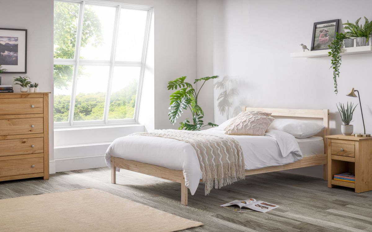 Unfinished pine double bed in natural wood finish, featuring a simple design with a low headboard, accompanied by a bedside table and decorative plants in a bright room.