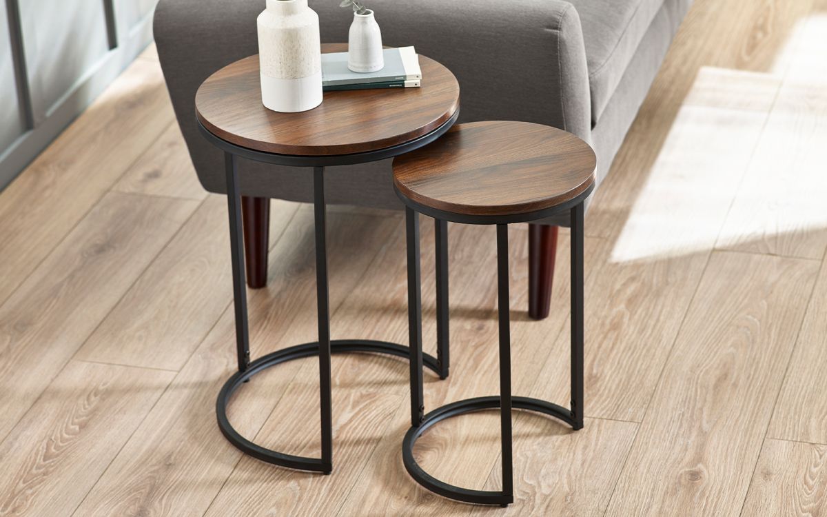 Round nesting side tables in walnut finish with sleek black metal legs, featuring a modern design, set beside a gray sofa on a light wooden floor.