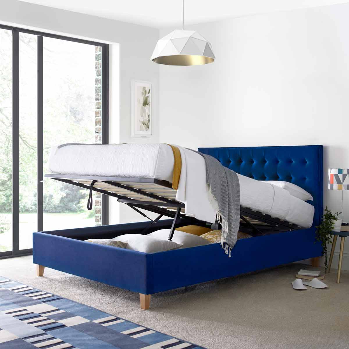 Kingham blue velvet ottoman bed with a tufted headboard, featuring a lift-up mattress for storage, set against a bright room with large windows and modern decor.