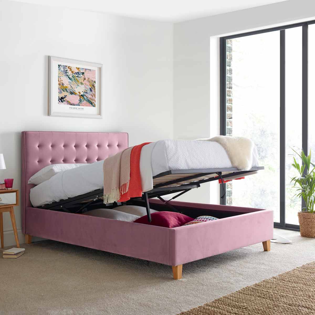 Kingham pink velvet ottoman bed with a tufted headboard, featuring a lift-up mattress revealing storage space beneath, set in a bright room with large windows.