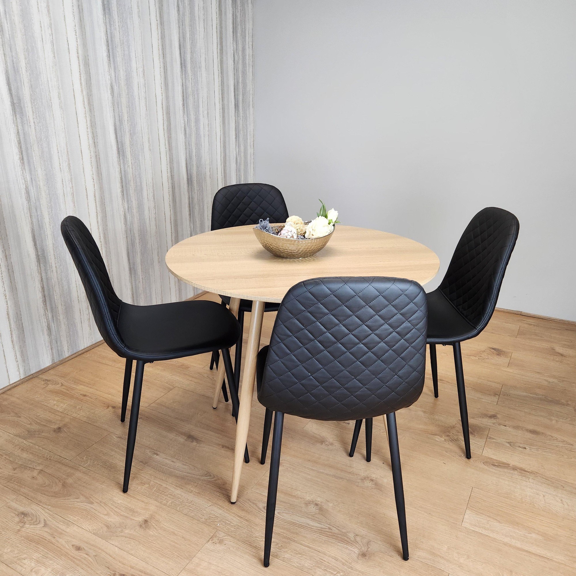 Round oak effect dining table with four black faux leather padded chairs, featuring a quilted design and slender black legs, set against a light wood floor and textured wall.