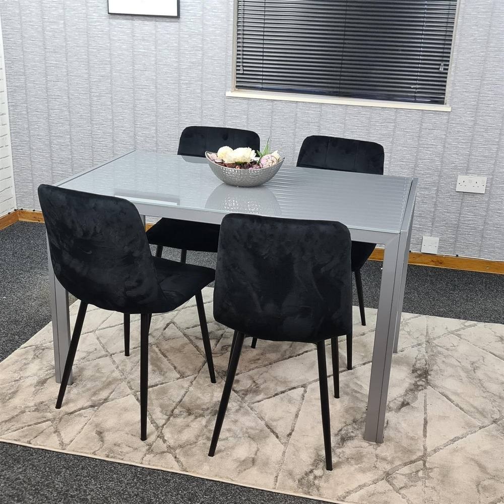 Grey dining table set featuring a sleek glass top and four black velvet chairs with slender black legs, arranged on a light-colored rug in a modern interior.