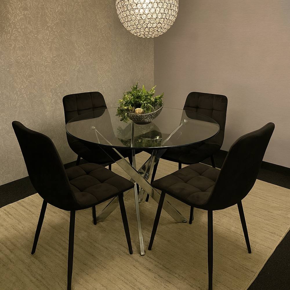 Round glass dining table with a sleek chrome base, surrounded by four black velvet chairs with tufted backs, set on a light-colored rug under a decorative pendant light.