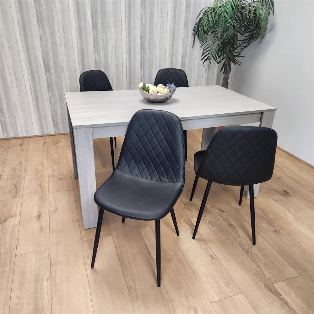 Grey wood dining table paired with four black leather chairs featuring diamond stitching, set on a light wooden floor with a decorative bowl of fruit on the table.