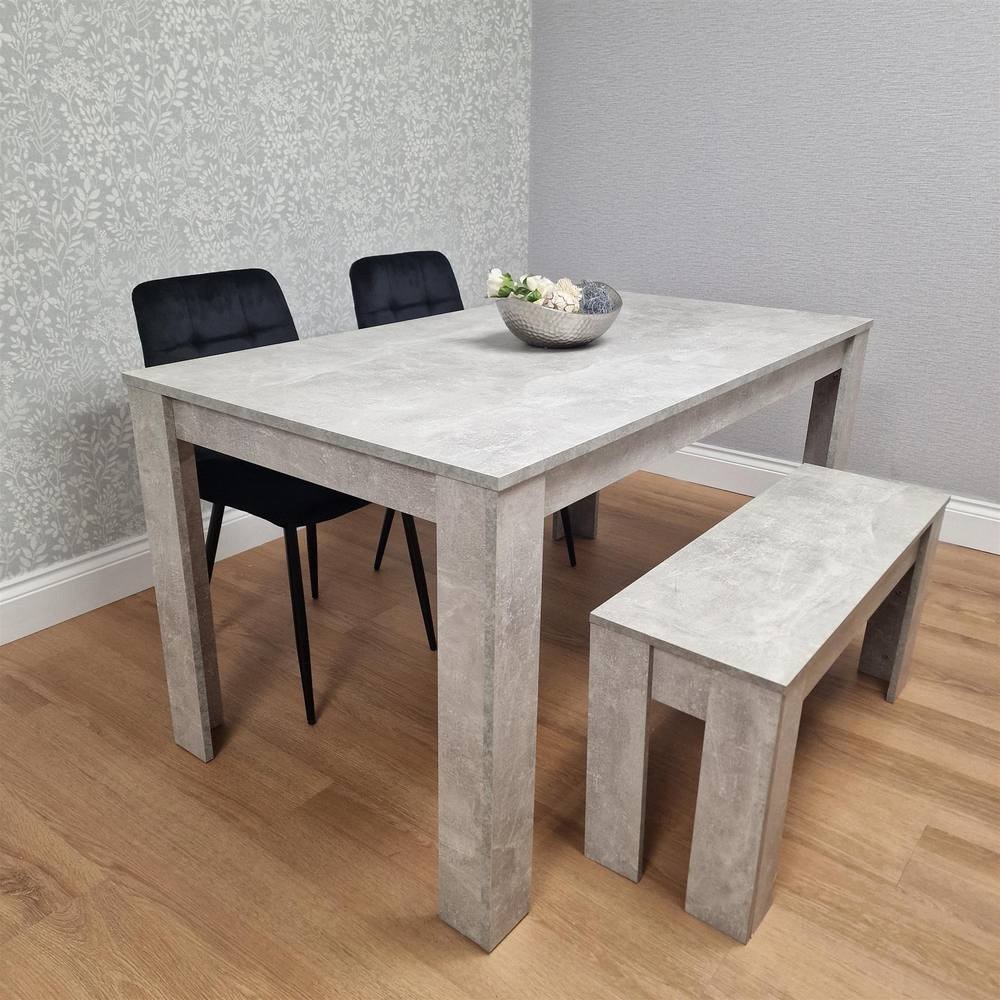 Grey concrete dining table set featuring two black upholstered chairs and a matching bench, all set against a light floral-patterned wall.