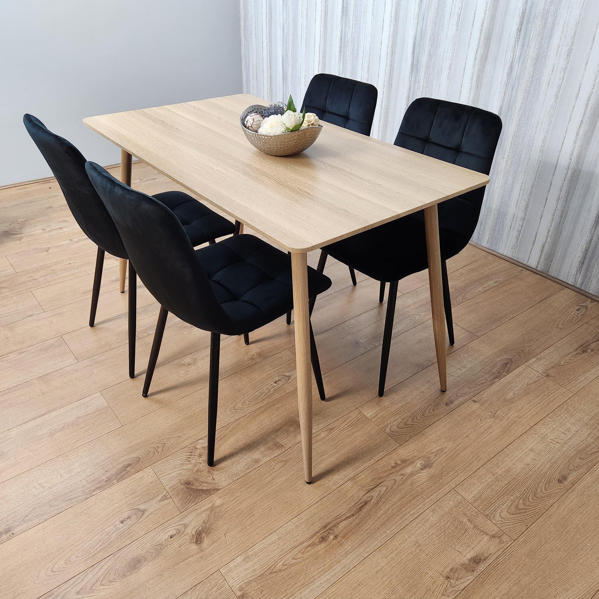 Rectangle oak effect dining table with four black velvet tufted chairs, featuring wooden legs, set against a light grey wall and wooden floor.
