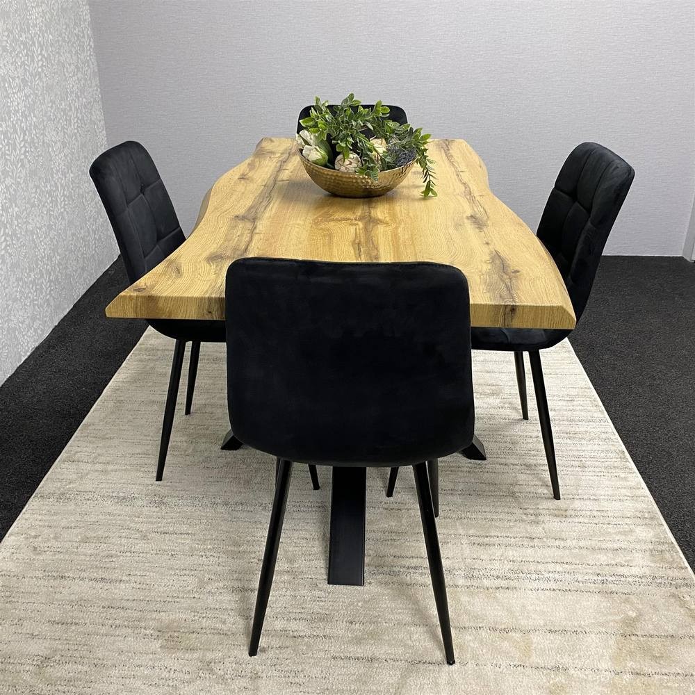 Stylish brown wooden dining table with a natural edge, accompanied by four black velvet chairs, set on a light-colored rug with a decorative bowl of greenery.