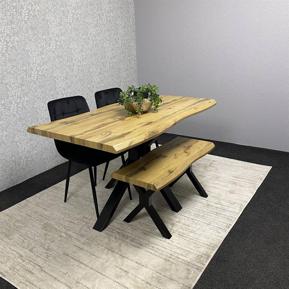 Kosy Koala wooden dining table in brown with a natural edge, accompanied by two black chairs and a matching bench, set on a light rug against a textured wall.