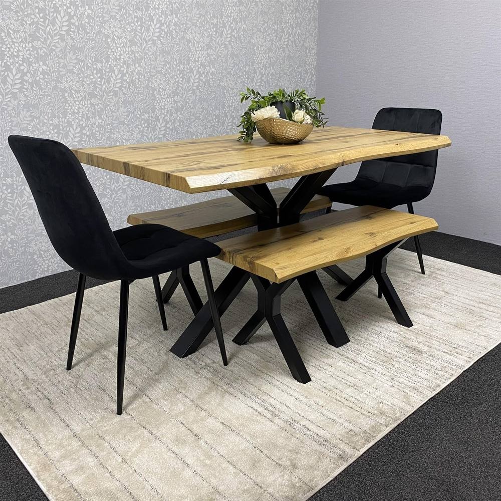 Stylish wooden dining table with a natural finish, black metal base, accompanied by a matching bench and two black velvet chairs, set against a light floral patterned wall.