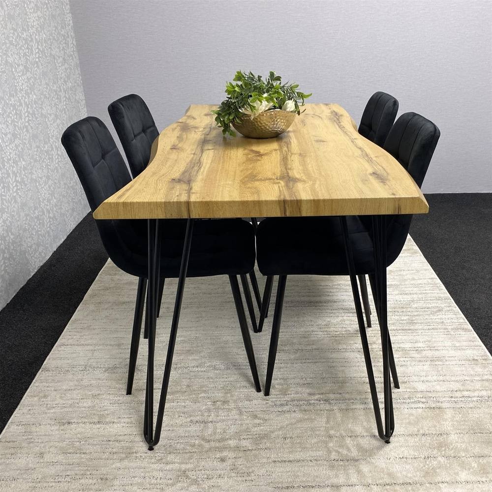 Brown wooden dining table with a natural finish, featuring four black upholstered chairs with slender black metal legs, set on a light-colored rug. A decorative bowl with greenery is on the table.