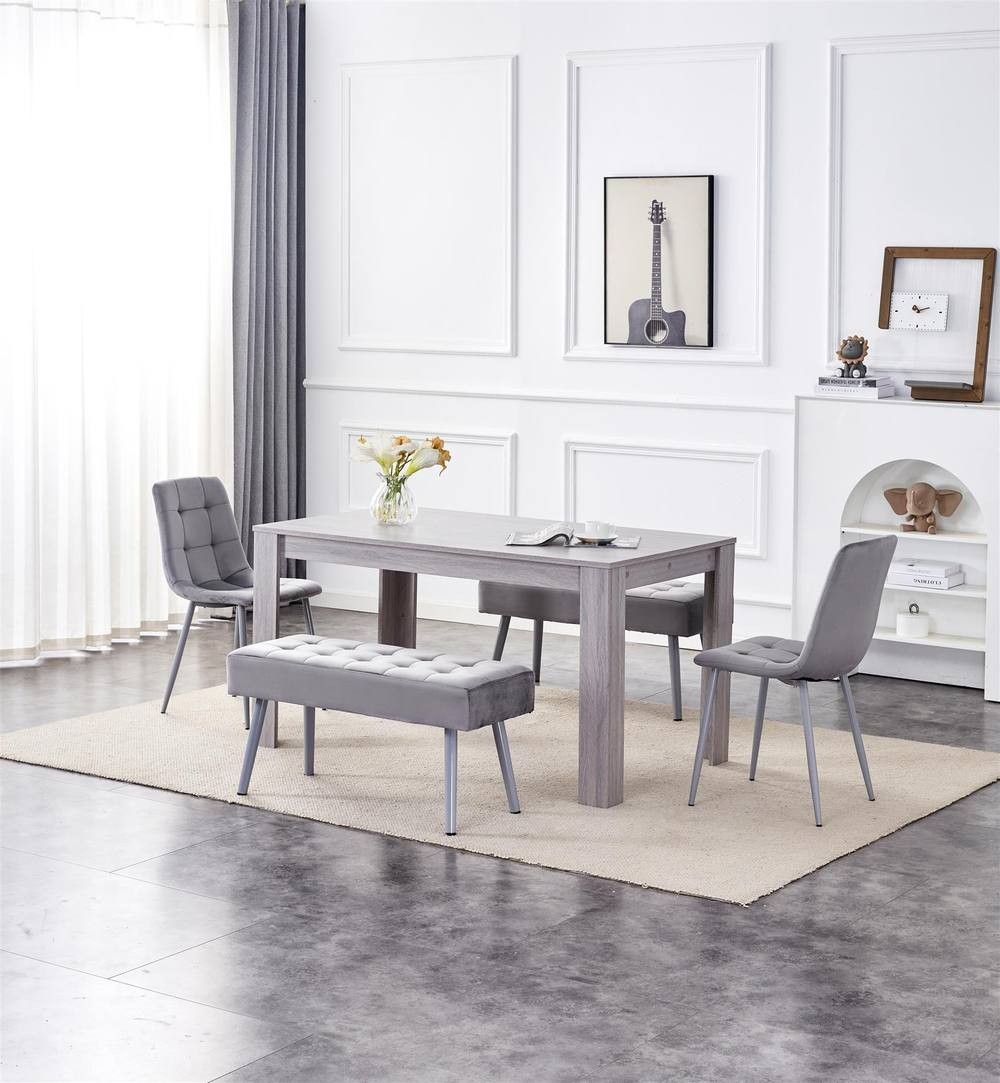 Grey wood dining table with two grey velvet chairs and two matching benches, set on a light rug in a bright room with white walls and natural light.