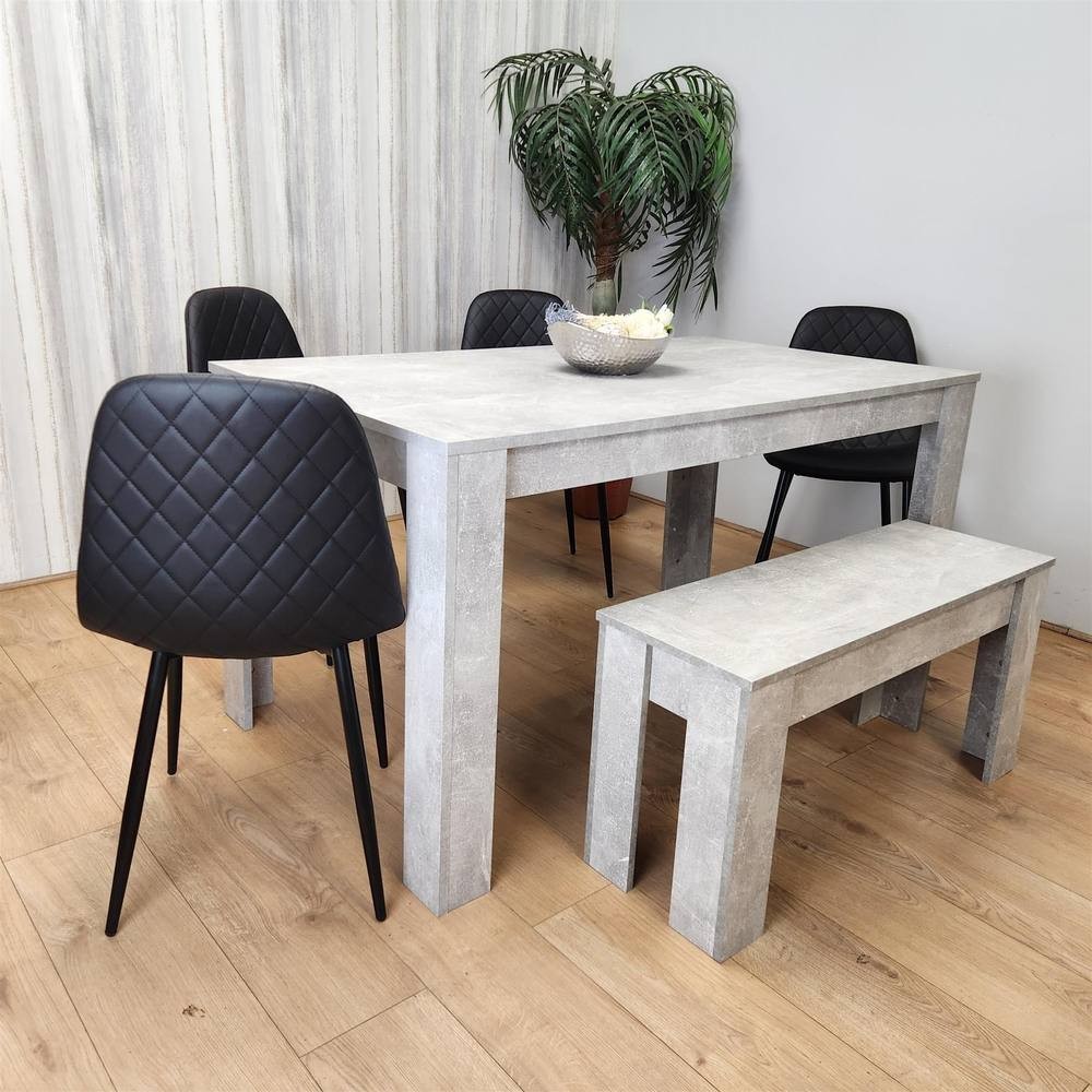 Stone grey wood dining table with a matching bench and four black leather chairs featuring diamond quilted patterns, set against a light wooden floor and a potted plant backdrop.