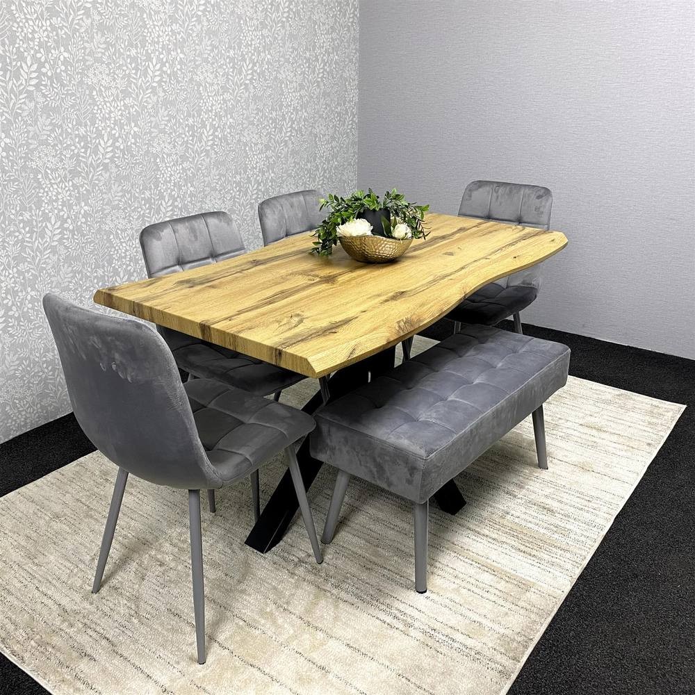 Kosy Koala wooden dining table with a natural finish, accompanied by two grey velvet chairs and a padded bench, set on a light rug against a textured wall.