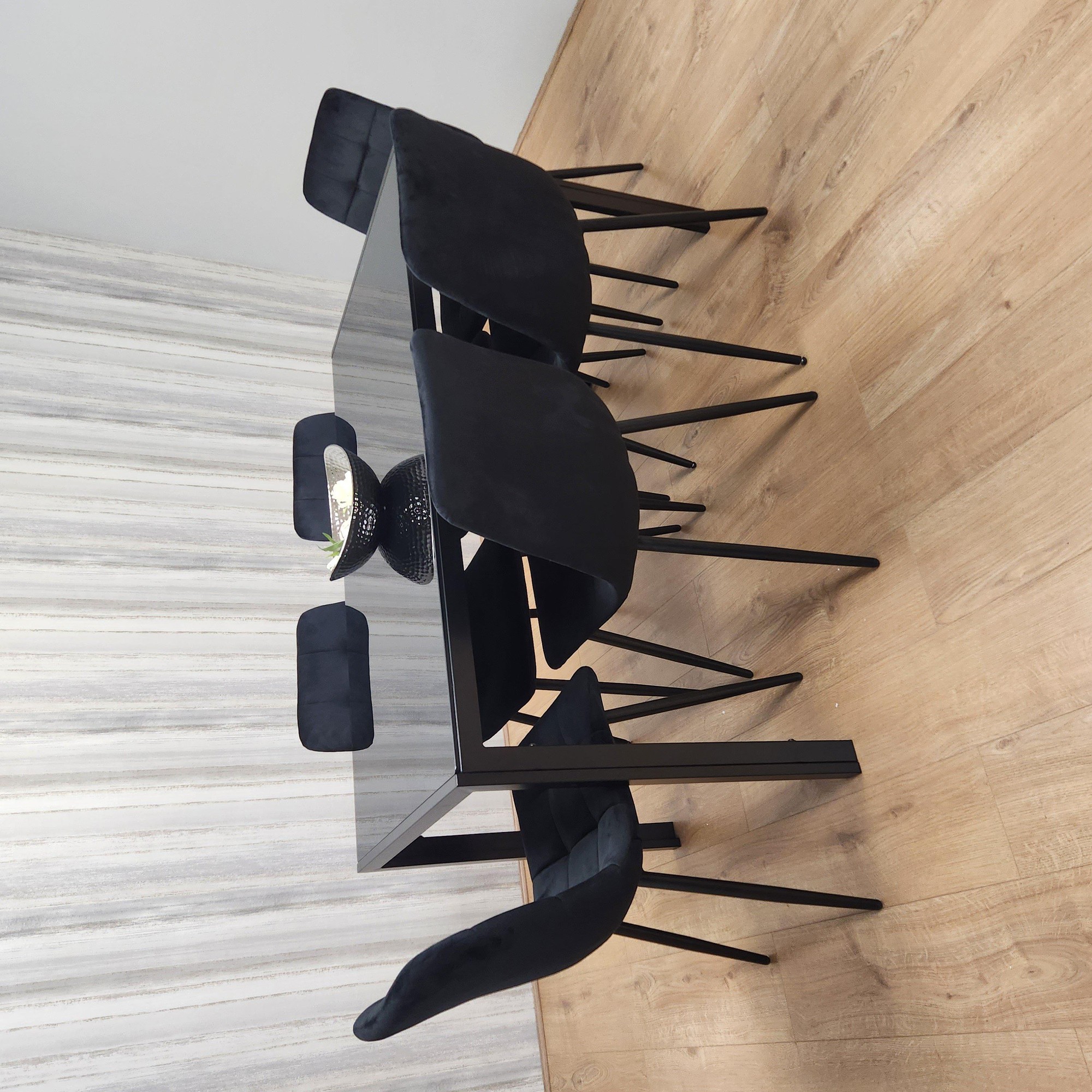 Black kitchen dining table with a sleek glass top and six black tufted velvet chairs featuring metal legs, set against a light wood floor and textured wall.