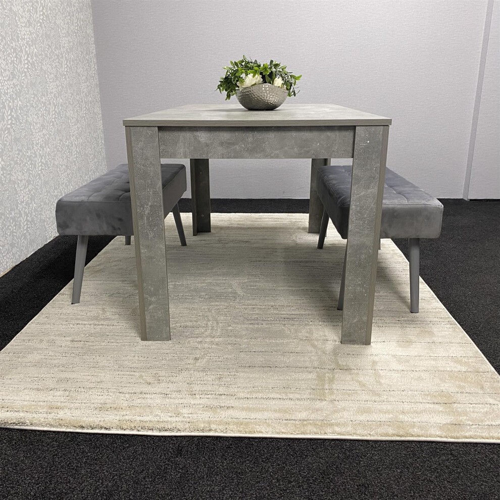 Kosy Koala wooden dining table in brown with two grey velvet chairs and padded benches, set on a light-colored rug in a minimalist dining room.
