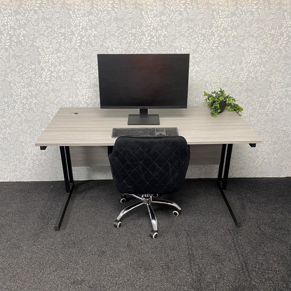 Grey L-shaped office desk with a sleek finish, accompanied by a grey velvet upholstered chair on wheels, featuring storage drawers and a small potted plant on the desk.