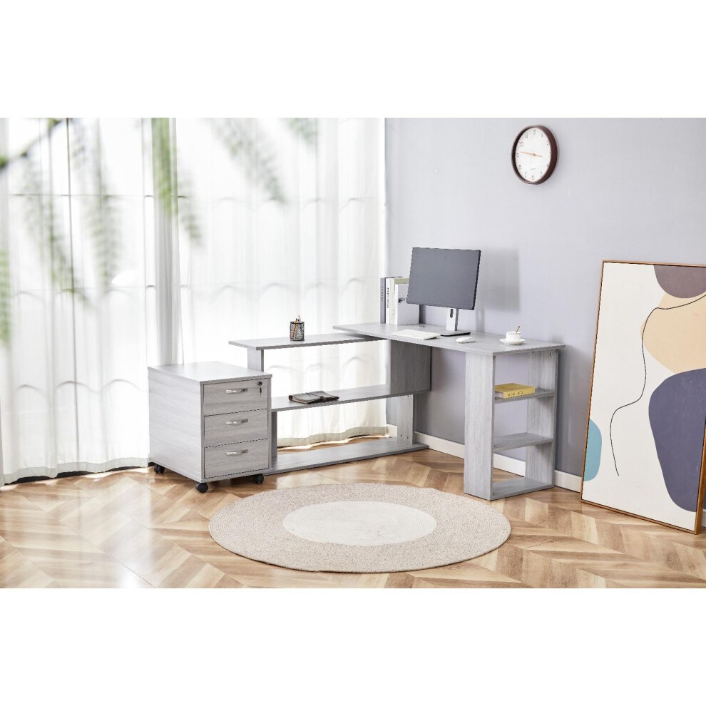 Grey L-shaped office desk made of wood, featuring a mobile storage drawer unit, open shelving, and a modern design, set in a bright room with a round rug and minimalist decor.