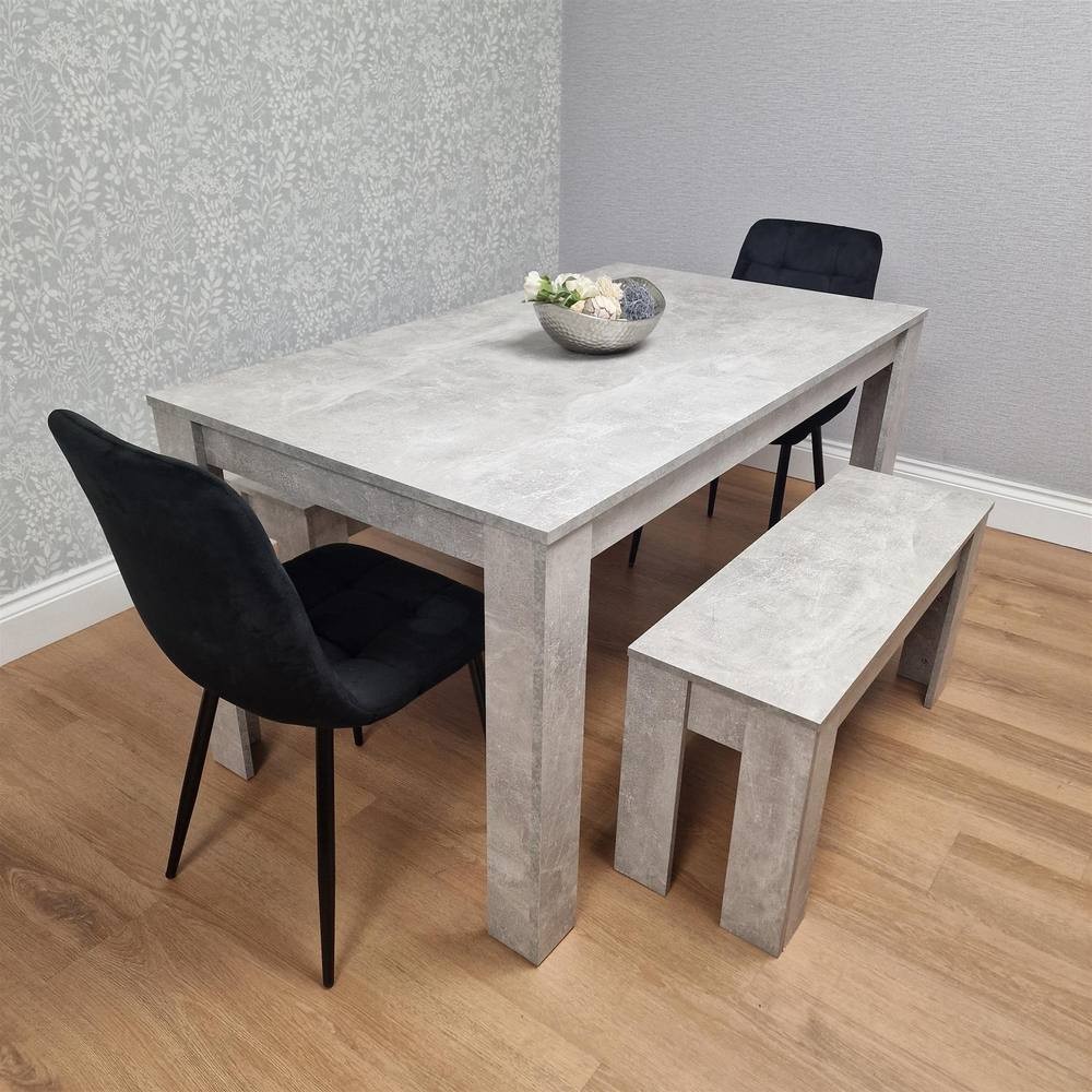 Modern dining set featuring a stone grey rectangular table, two black velvet chairs with sleek legs, and a matching bench, set against a patterned wall and wooden floor.