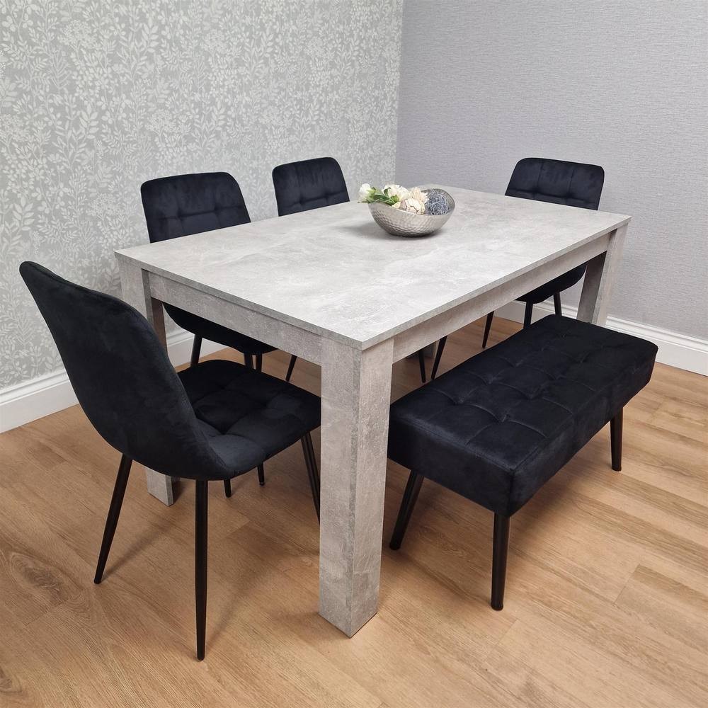 Modern grey dining table with a textured finish, accompanied by four black velvet chairs and a black velvet bench, all set against a light floral-patterned wall.