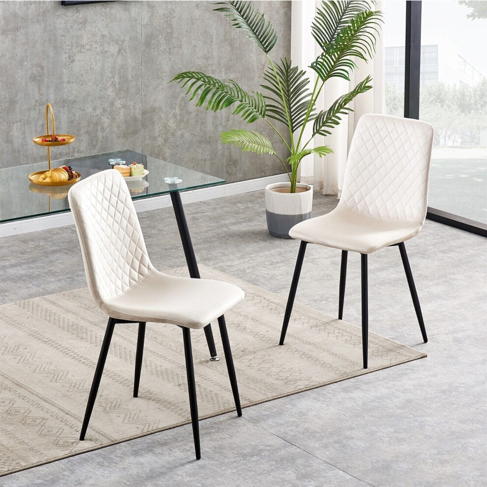Beige and cream velvet dining chairs with diamond quilted pattern, featuring sleek black metal legs, set against a modern interior with a glass table and greenery.