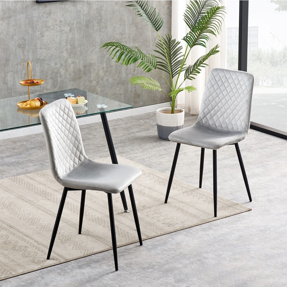 Light grey velvet dining chairs with diamond quilted pattern, black metal legs, set of two, placed on a neutral-toned rug in a modern interior with a glass table and greenery.