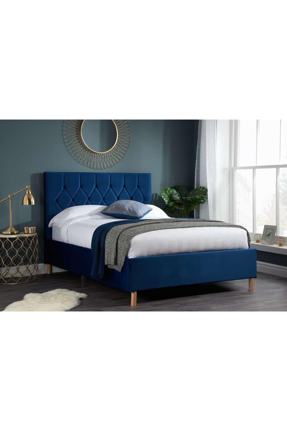 Small double bed in deep blue velvet with a tufted headboard, complemented by wooden legs, styled with a white duvet and decorative pillows against a modern room backdrop.