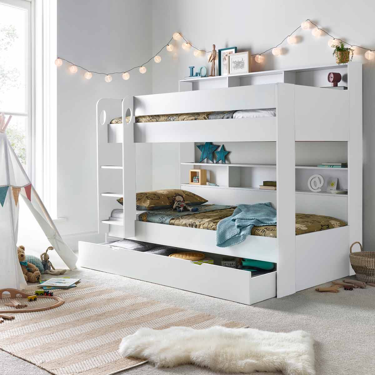 White wooden bunk bed with a lower storage drawer, featuring a ladder, shelves for books, and two single mattresses, set in a cozy room with decorative fairy lights.