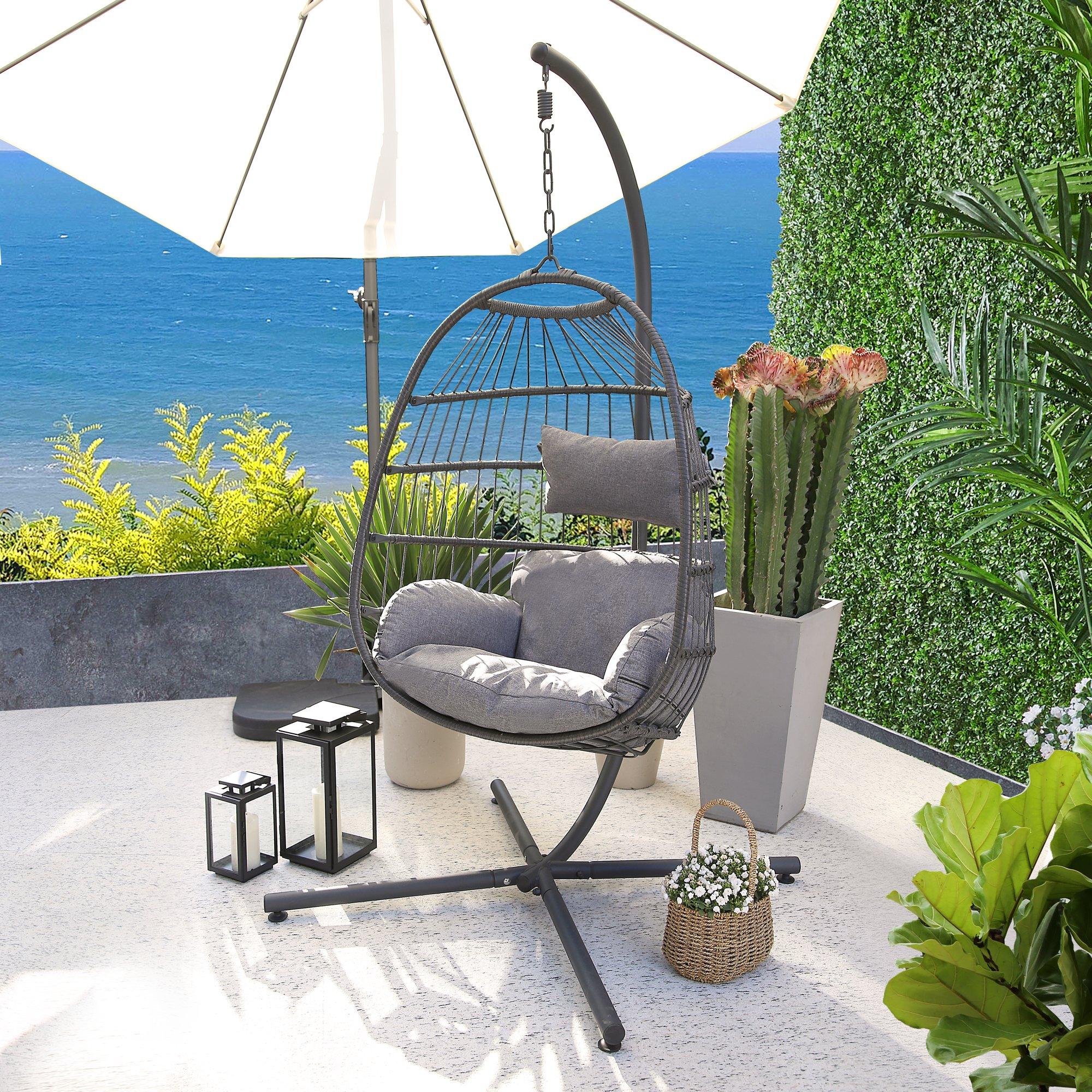 Azura Hanging Egg Chair features a large, woven design in deep grey with plush cushions, suspended from a sturdy black metal frame, set against a scenic outdoor backdrop.