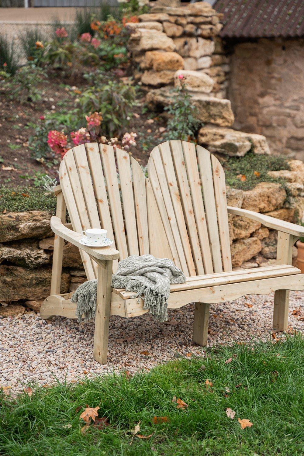Natural wood double Adirondack garden bench with a curved backrest, featuring a cozy throw blanket and a teacup on the armrest, set against a stone backdrop and greenery.