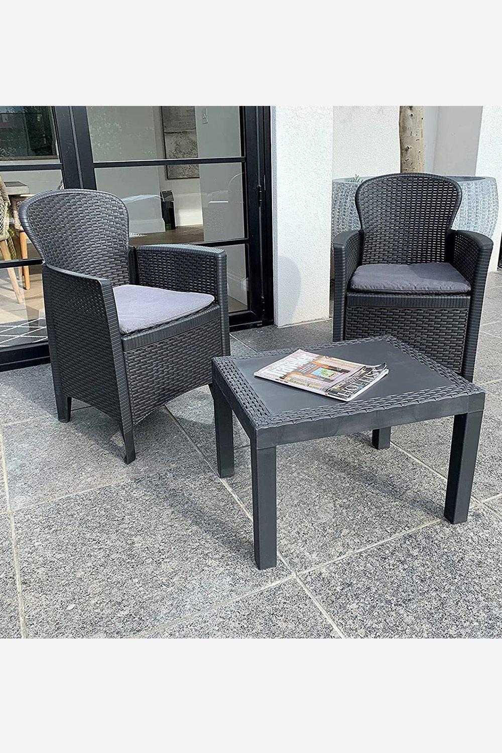 Two black rattan effect chairs with gray cushions and a matching square table, set on a stone patio, ideal for outdoor dining or relaxation.