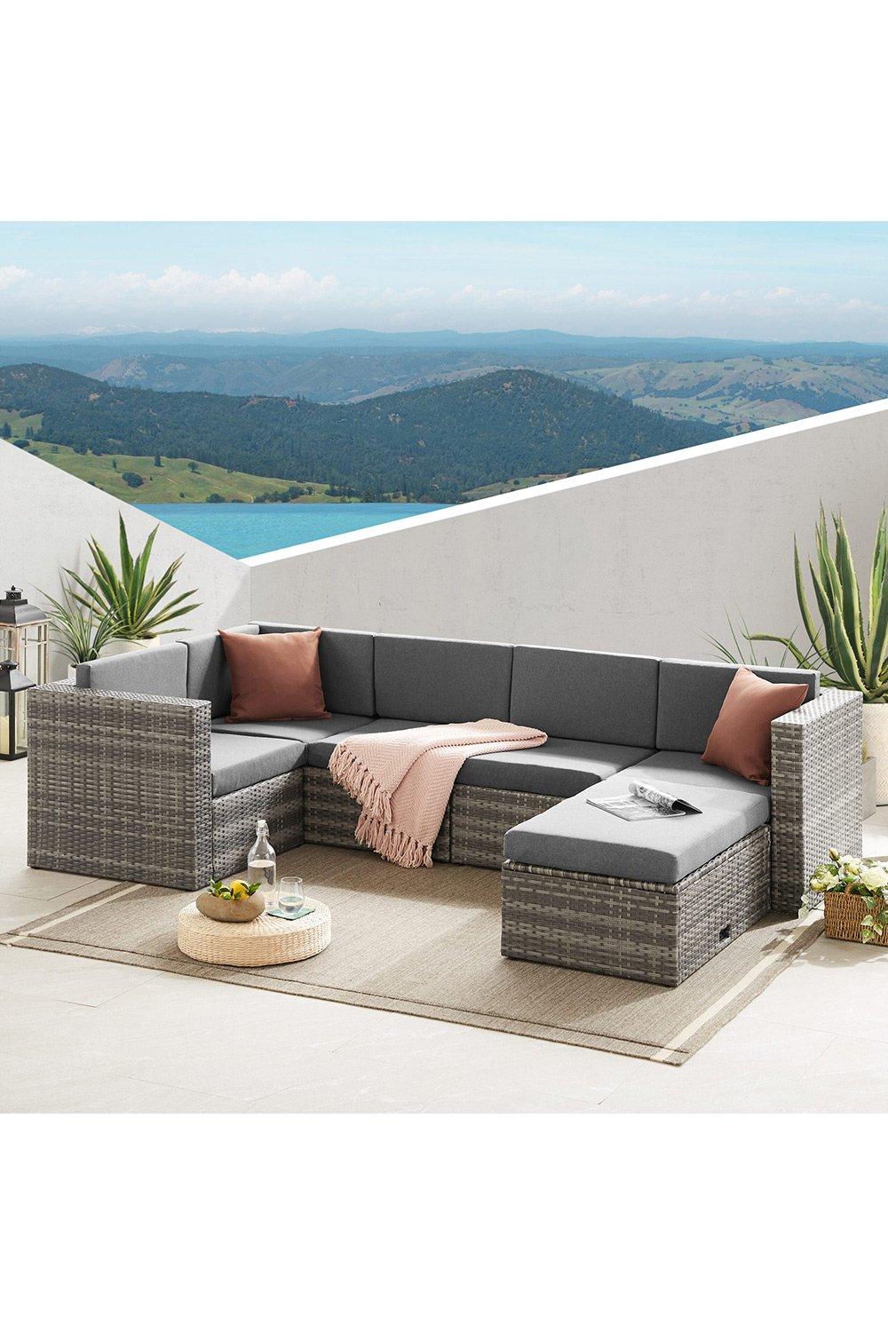 Grey rattan corner sofa set with six seats, featuring plush grey cushions, a light pink throw, and a modern design, set against a scenic outdoor backdrop.