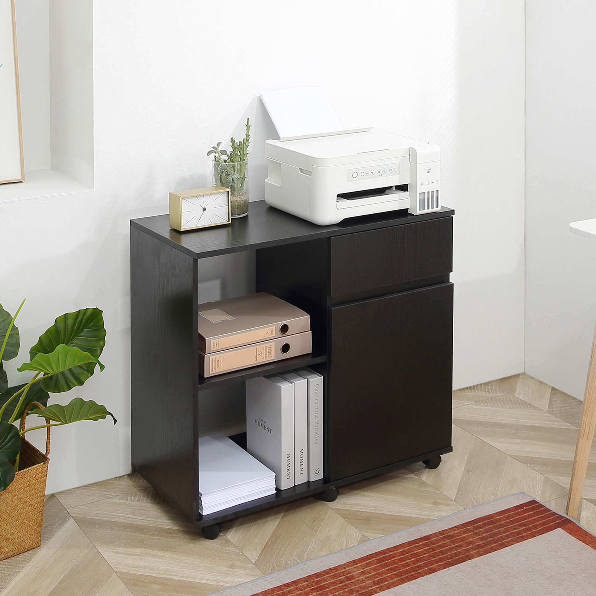 Black wooden filing cabinet with open storage shelves, an easy drawer, and a printer on top, positioned next to a potted plant in a bright room.