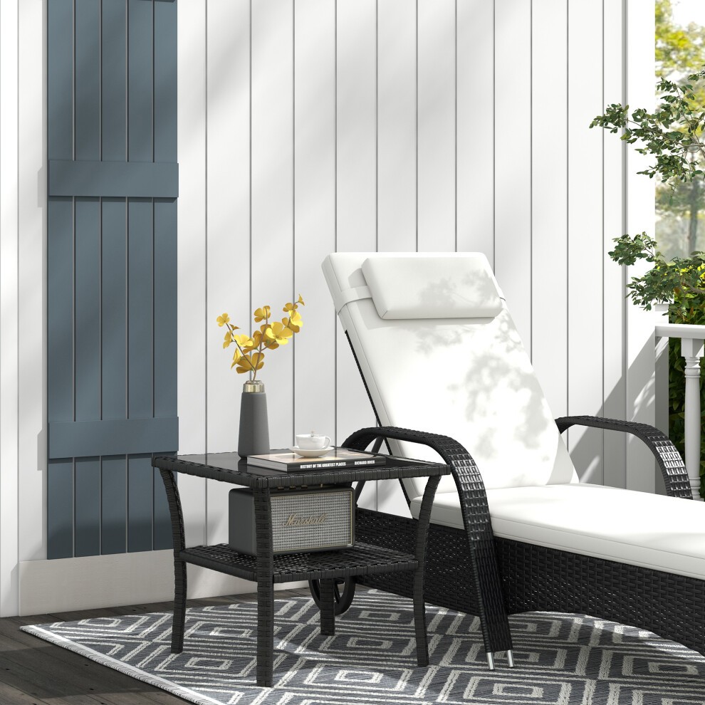 Black PE rattan coffee table with a two-tier design and a glass top, positioned beside a white lounge chair, featuring a decorative vase with yellow flowers.