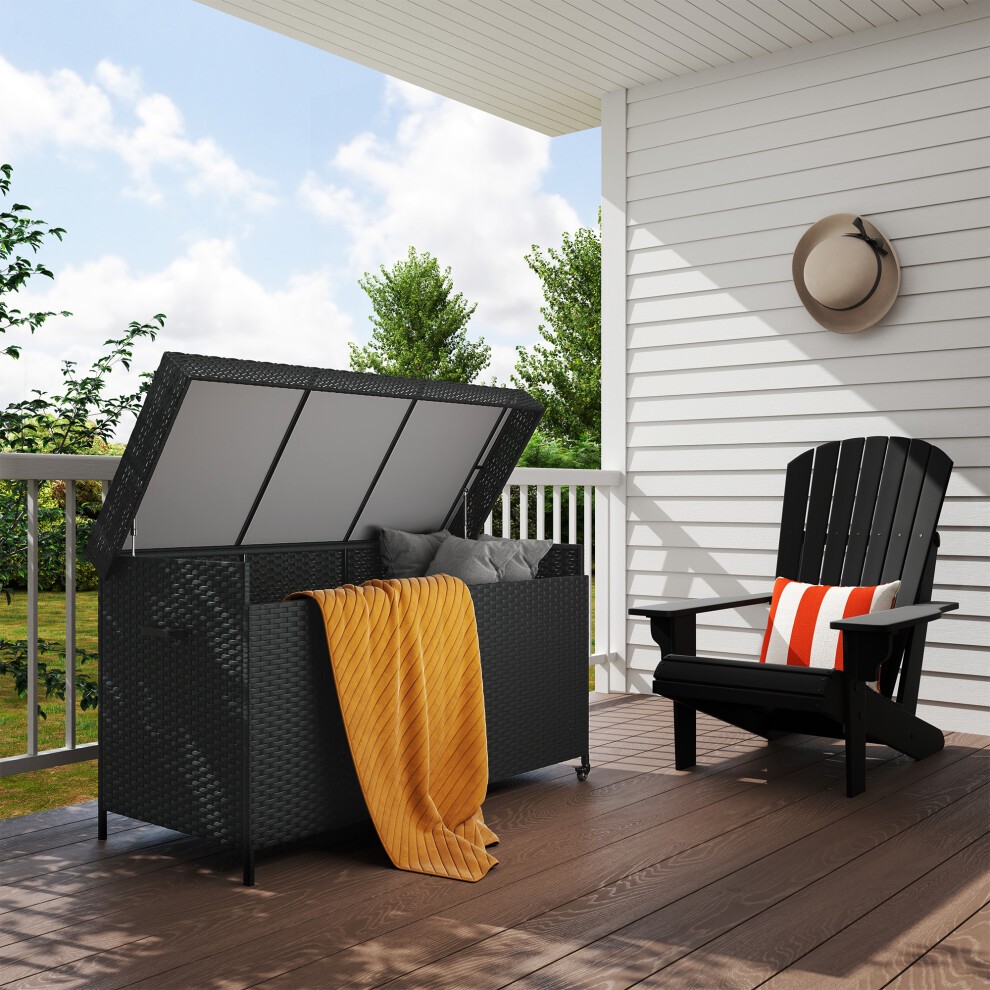 Black rattan garden storage box with a gas lift lid and wheels, featuring a cozy orange blanket inside, placed on a wooden deck beside a black adirondack chair.